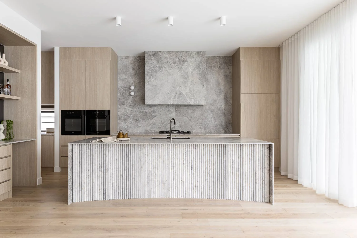 Luxury-Modern-Kitchen-Design-Marble-Fluted-Kitchen-Island-Oak-Cabinetry-White-Curtains-Brushed-Gunmetal-Tapware